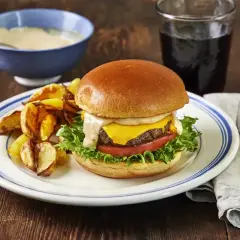 En saftig hamburgare med ost, sallad och tomat, serverad med klyftpotatis och en skål med krämig hamburgerdressing.