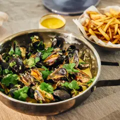 En stor kastrull med ångande moules frites, garnerad med persilja, bredvid en skål med gyllene pommes frites.