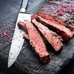 Skivat flap steak med rosa salt på en sotad träskärbräda, bredvid en kniv.