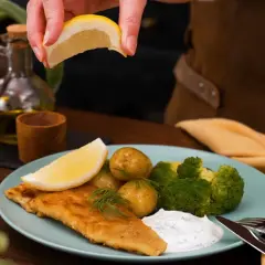 En saftig panerad fisk serveras med potatis, broccoli och en klick vit sås. En citronskiva pressas över fisken för extra smak.