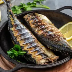 Två hela, grillade makrillar ligger i en gjutjärnspanna med citronklyftor och persilja.