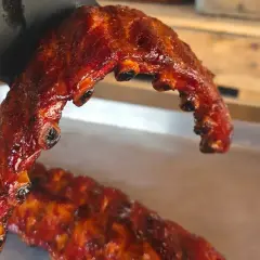 En hand i svart handske håller upp saftiga, glaserade grillade revben. En annan rad revben ligger på en bricka.