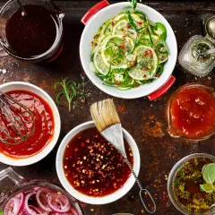 Olika såser och ingredienser för ett chilisås recept. En visp i en skål med röd sås, en pensel i en skål med mörk sås, och skivade lime och jalapeño.