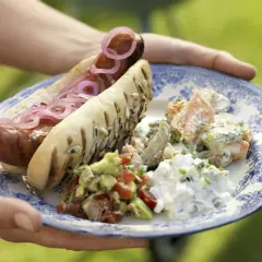 En tallrik med en grillad korv i ett bröd med rödlök, guacamole, potatissallad och äggsallad. Ett perfekt korv recept för sommaren.