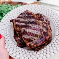 En hand med rött nagellack håller upp en tallrik med saftig biff tagliata, grillad till perfektion. Gröna blad syns i bakgrunden.