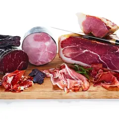 En träskärbräda med olika sorters charkuterier, inklusive en stor bit lufttorkad skinka, skivor av prosciutto och en rund, kokt skinka.