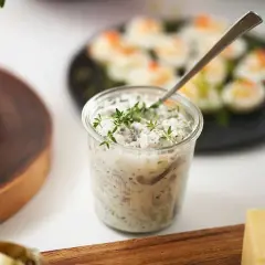 En burk med krämig wasabi sill, toppad med färska örter. I bakgrunden syns ägghalvor och en bit bröd.