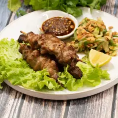 Grillade spett med fläsk, kända som moo ping, serveras med en fräsch gurksallad, dippsås och citronklyfta på en bädd av sallad.