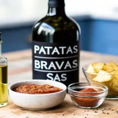 En flaska patatas bravas-sås, olivolja, två skålar med sås och en skål med klyftpotatis. Perfekt för en mysig kväll med grillad potatis.
