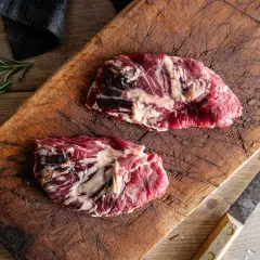 Två råa spider steak-biffar på en sliten träskärbräda, med rosmarin och en kniv.