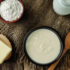 En skål med krämig vit sås, bredvid smör, mjöl och en mjölkburk. Ingredienser för en klassisk vit sås.
