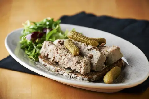 En tallrik med skivad leverpastej på mörkt bröd, toppad med cornichoner och en sallad. Perfekt för ett klassiskt leverpastej recept.