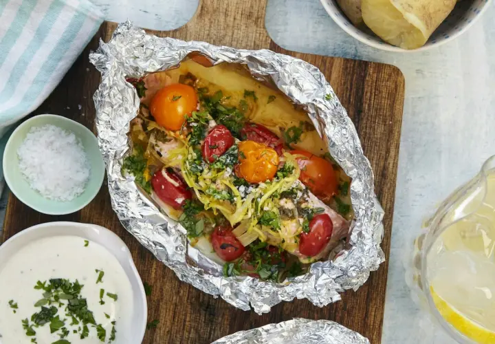 En saftig laxsida i foliepaket med tomater och örter, redo att grilla. Serveras med potatis och en krämig sås.