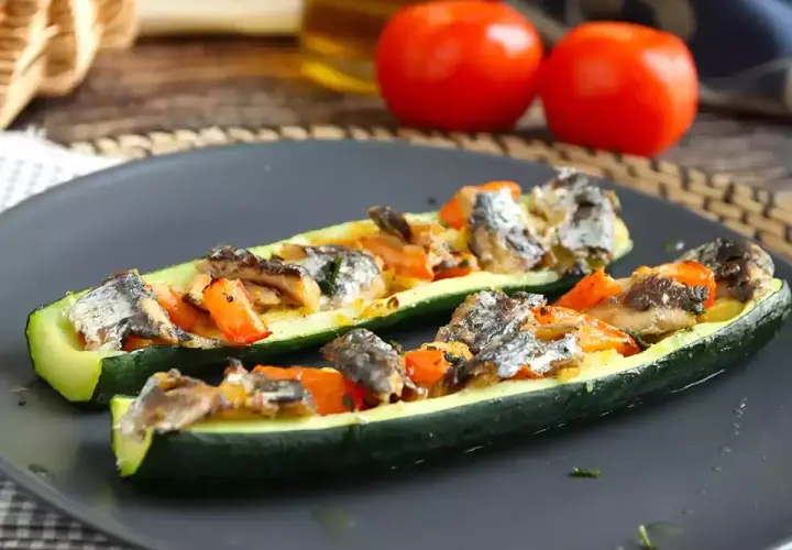 Två halvor av fylld zucchini med fisk och grönsaker på en mörk tallrik.