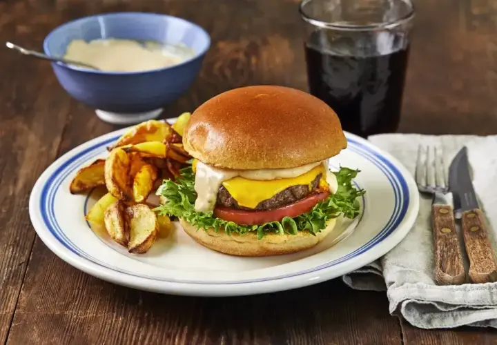 En saftig hamburgare med ost, sallad och tomat, serverad med klyftpotatis och en skål med krämig hamburgerdressing.