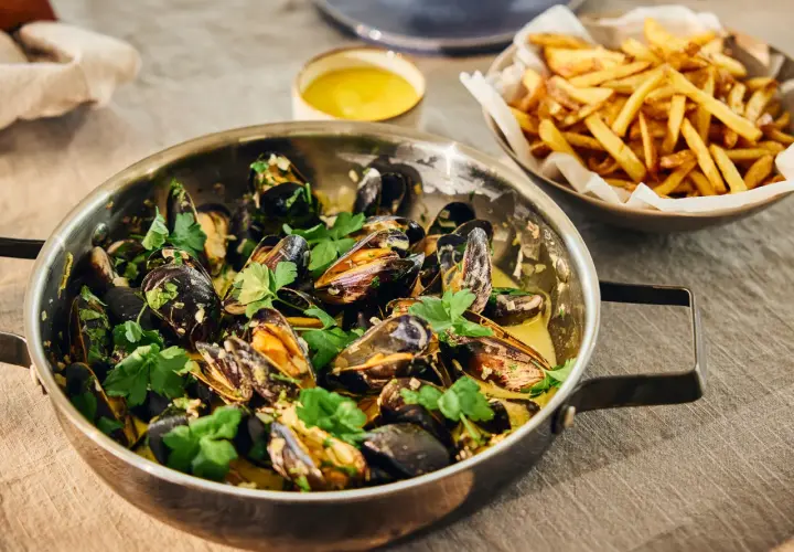 En stor kastrull med ångande moules frites, garnerad med persilja, bredvid en skål med gyllene pommes frites.