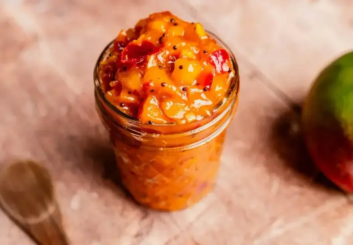 En burk med mango chutney, perfekt för ditt nästa chutney recept. En hel mango och en träsked syns också.