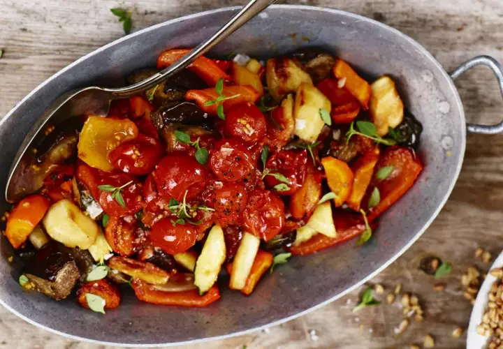 En varm ugnsbakad grönsaksblandning med körsbärstomater, paprika och aubergine, serverad i en metallskål med en sked. En härlig rostad tomatsås gör rätten extra smakrik.