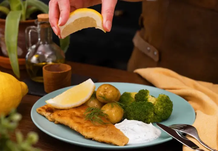 En saftig panerad fisk serveras med potatis, broccoli och en klick vit sås. En citronskiva pressas över fisken för extra smak.