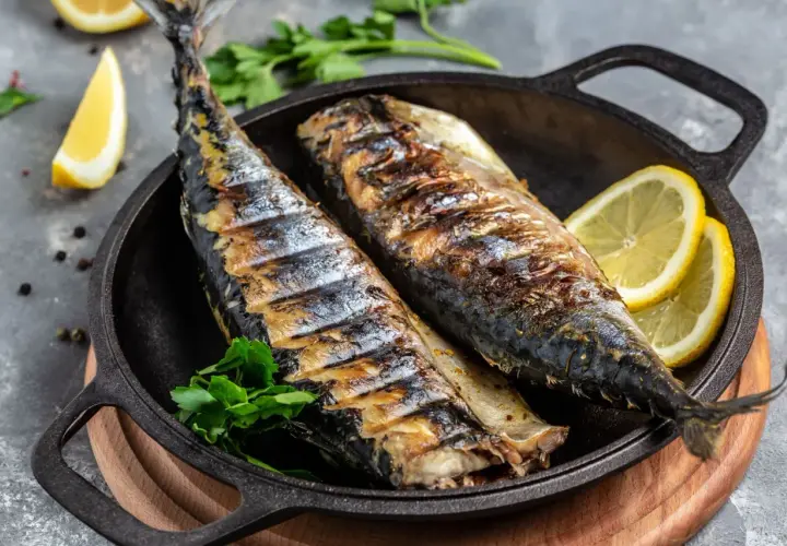 Två hela, grillade makrillar ligger i en gjutjärnspanna med citronklyftor och persilja.