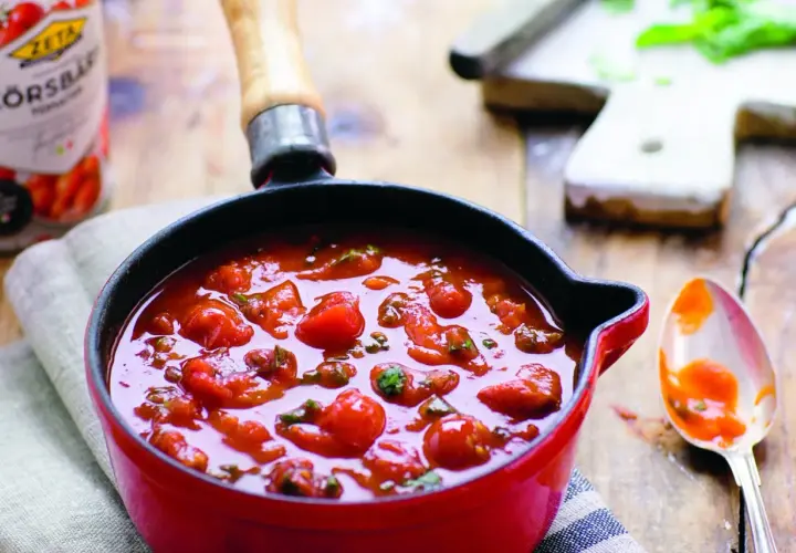 En röd kastrull fylld med en smakrik italiensk tomatsås med hela körsbärstomater och örter. En sked med sås ligger bredvid.