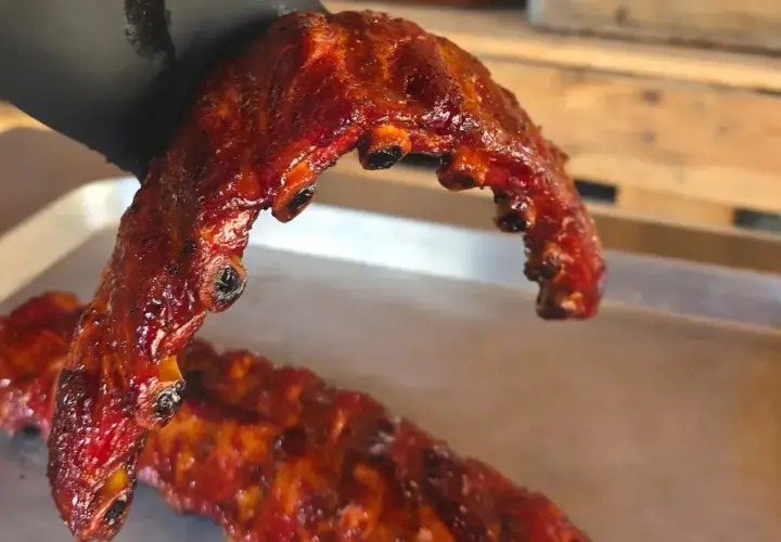 En hand i svart handske håller upp saftiga, glaserade grillade revben. En annan rad revben ligger på en bricka.