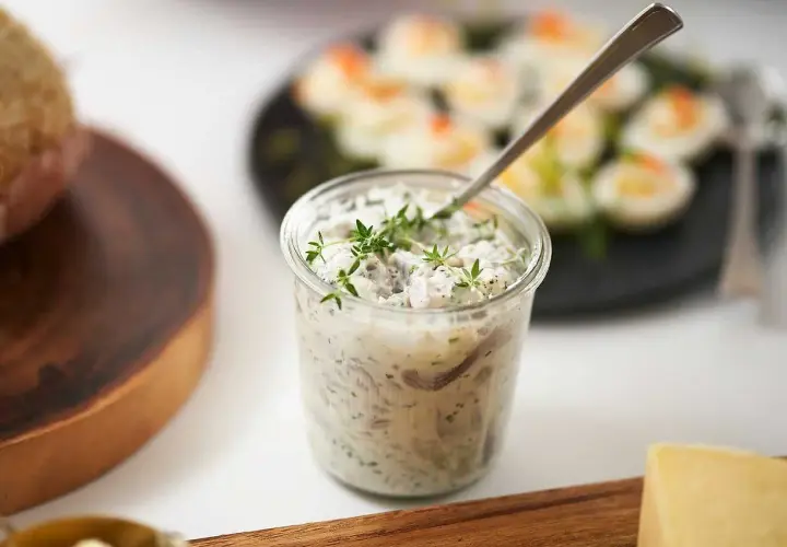 En burk med krämig wasabi sill, toppad med färska örter. I bakgrunden syns ägghalvor och en bit bröd.
