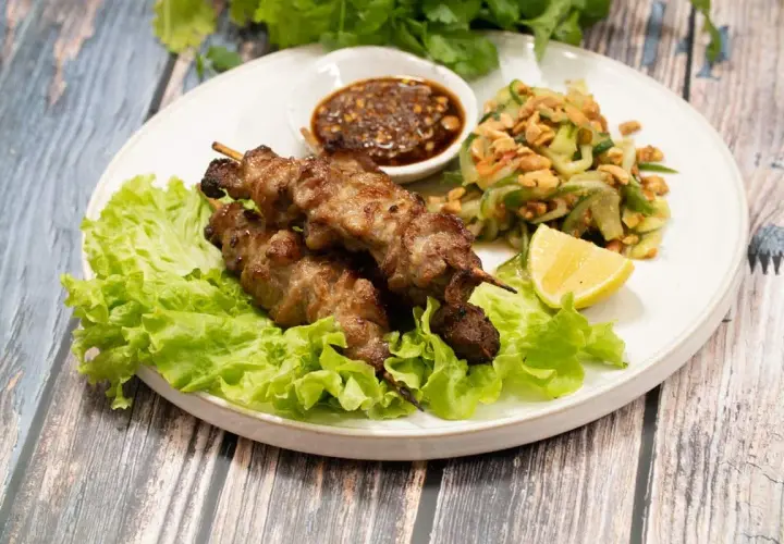 Grillade spett med fläsk, kända som moo ping, serveras med en fräsch gurksallad, dippsås och citronklyfta på en bädd av sallad.