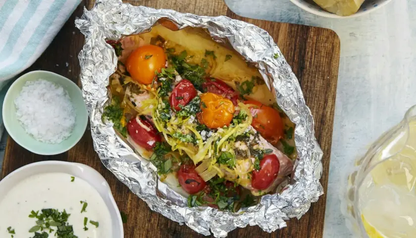 En saftig laxsida i foliepaket med tomater och örter, redo att grilla. Serveras med potatis och en krämig sås.