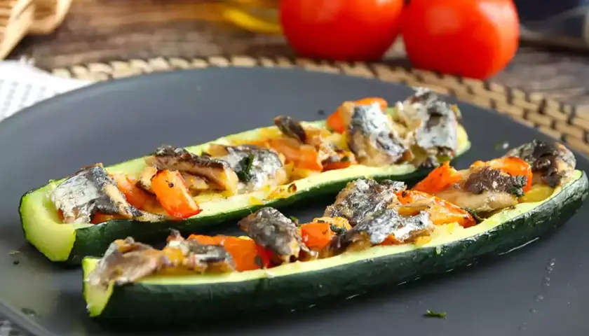Två halvor av fylld zucchini med fisk och grönsaker på en mörk tallrik.