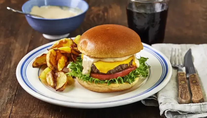 En saftig hamburgare med ost, sallad och tomat, serverad med klyftpotatis och en skål med krämig hamburgerdressing.
