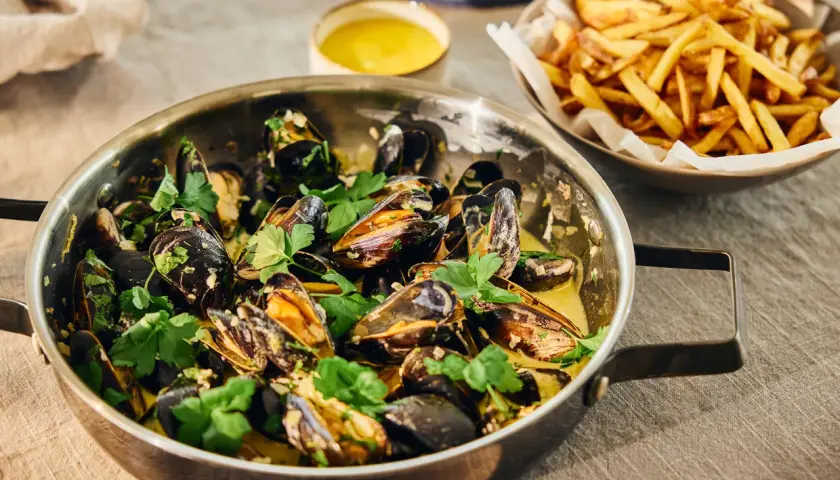 En stor kastrull med ångande moules frites, garnerad med persilja, bredvid en skål med gyllene pommes frites.