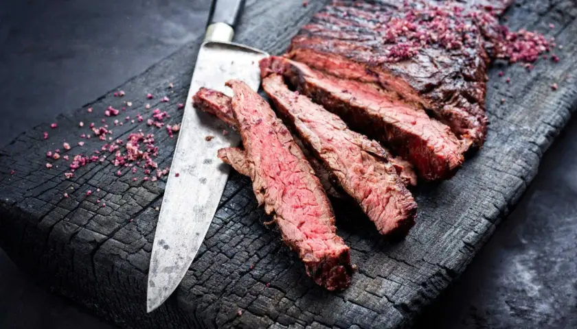 Skivat flap steak med rosa salt på en sotad träskärbräda, bredvid en kniv.