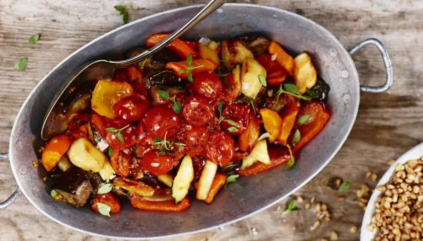 En varm ugnsbakad grönsaksblandning med körsbärstomater, paprika och aubergine, serverad i en metallskål med en sked. En härlig rostad tomatsås gör rätten extra smakrik.