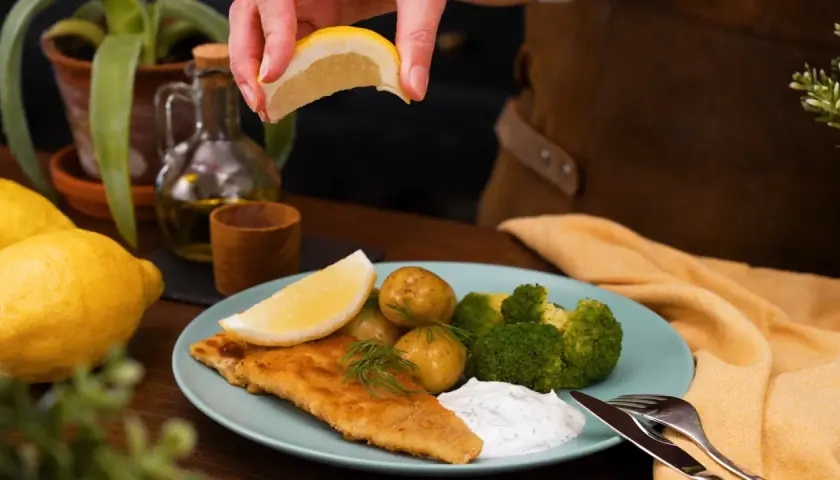 En saftig panerad fisk serveras med potatis, broccoli och en klick vit sås. En citronskiva pressas över fisken för extra smak.