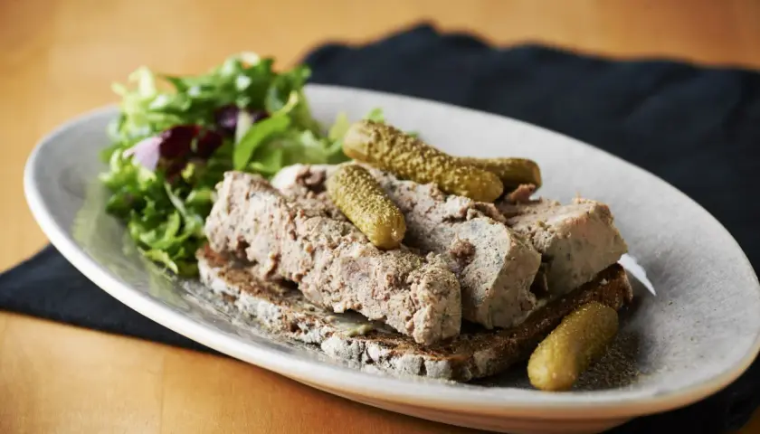 En tallrik med skivad leverpastej på mörkt bröd, toppad med cornichoner och en sallad. Perfekt för ett klassiskt leverpastej recept.