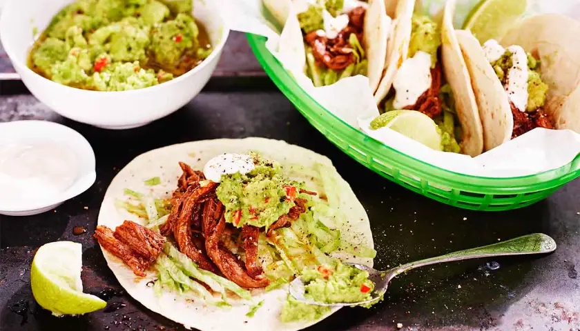 En läcker pulled beef taco med guacamole och strimlad sallad. En skål med mer guacamole och en limeklyfta syns också.