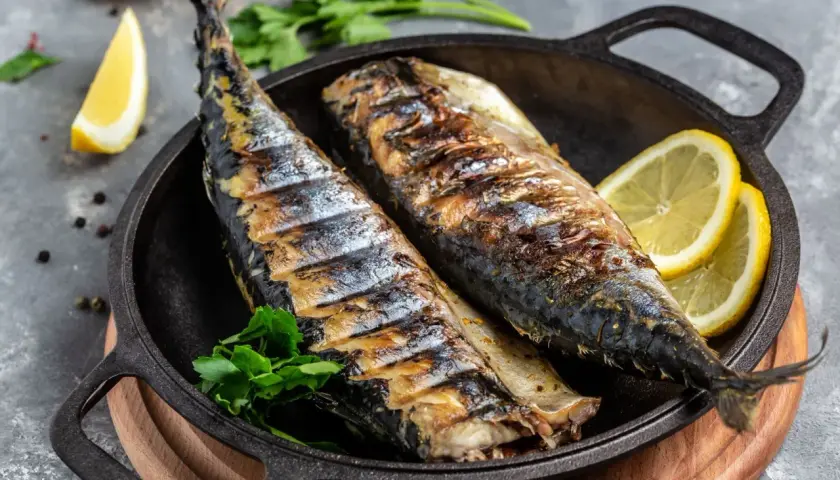 Två hela, grillade makrillar ligger i en gjutjärnspanna med citronklyftor och persilja.