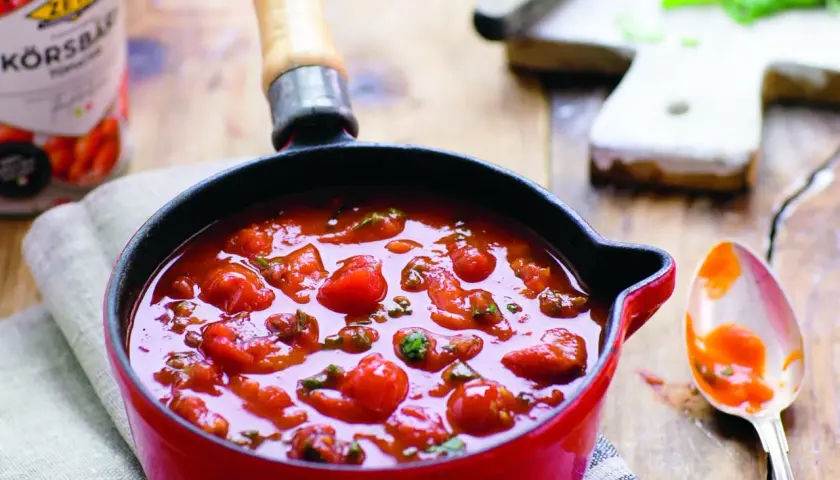En röd kastrull fylld med en smakrik italiensk tomatsås med hela körsbärstomater och örter. En sked med sås ligger bredvid.