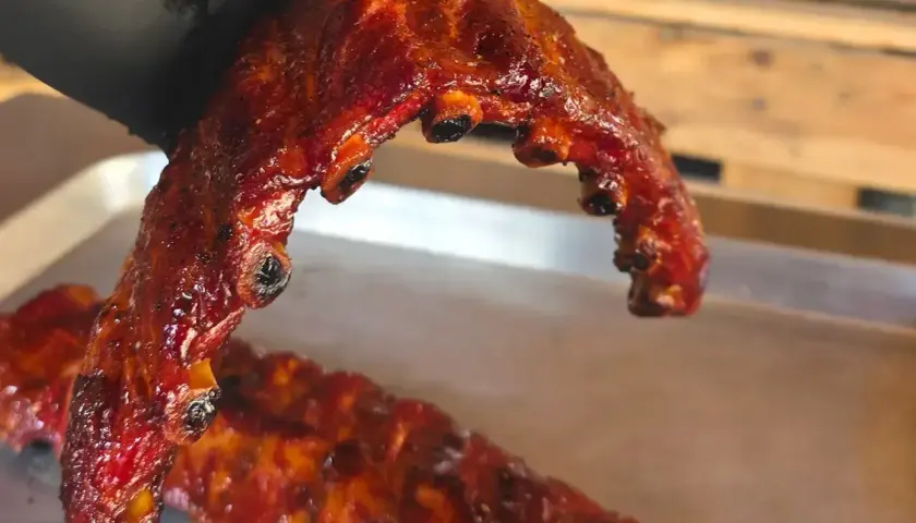En hand i svart handske håller upp saftiga, glaserade grillade revben. En annan rad revben ligger på en bricka.