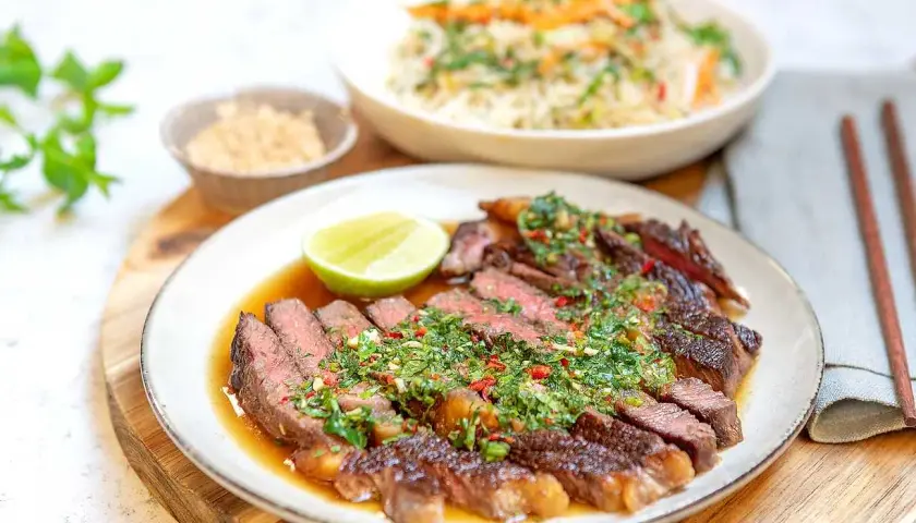 Skivad asiatisk entrecote med örtig sås, lime och en skål med ris.