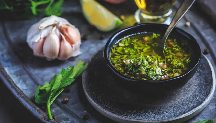 En skål med färsk chimichurri, redo att förhöja din nästa måltid. Ingredienser som vitlök, persilja och citron syns i närheten.