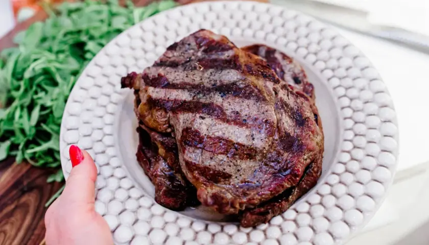 En hand med rött nagellack håller upp en tallrik med saftig biff tagliata, grillad till perfektion. Gröna blad syns i bakgrunden.