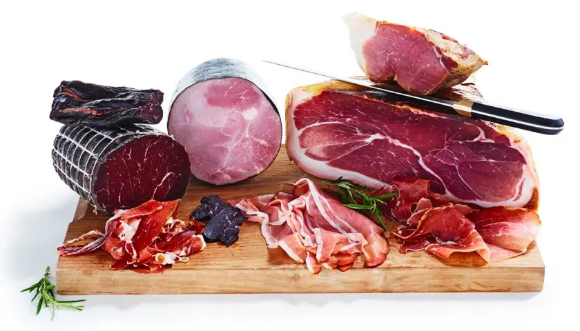 En träskärbräda med olika sorters charkuterier, inklusive en stor bit lufttorkad skinka, skivor av prosciutto och en rund, kokt skinka.