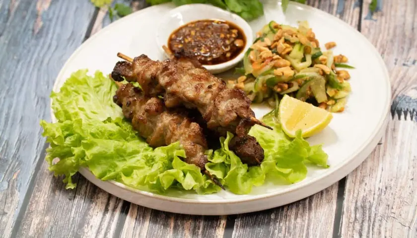 Grillade spett med fläsk, kända som moo ping, serveras med en fräsch gurksallad, dippsås och citronklyfta på en bädd av sallad.