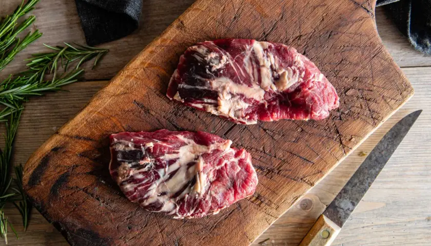Två råa spider steak-biffar på en sliten träskärbräda, med rosmarin och en kniv.