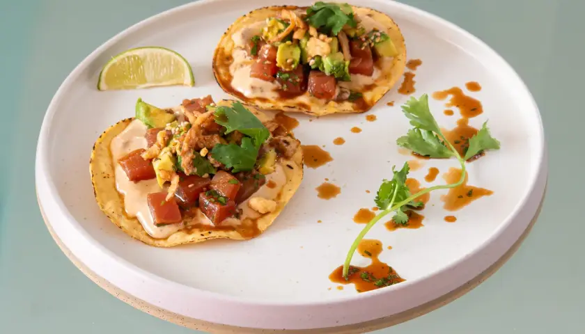 Två läckra tostadas med tonfisk tartar och avocado, toppade med koriander och en krämig sås. En limeklyfta ligger bredvid.
