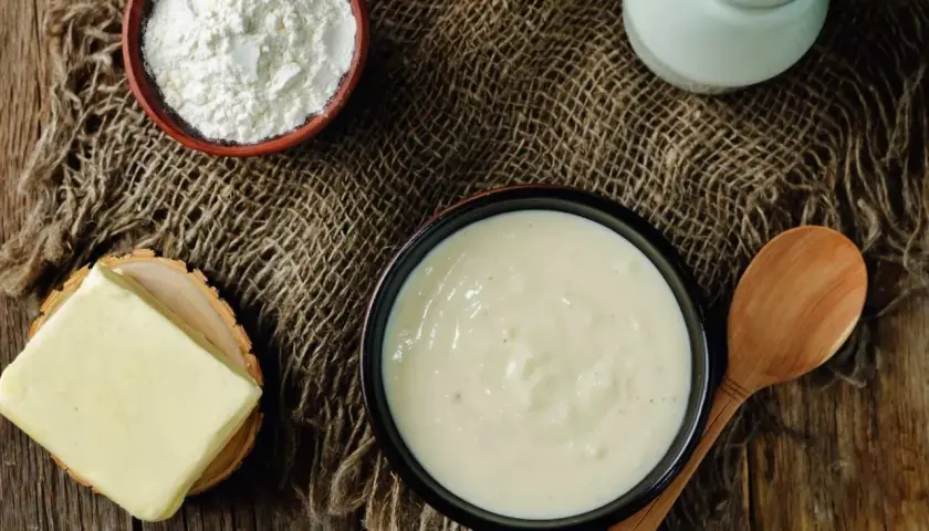 En skål med krämig vit sås, bredvid smör, mjöl och en mjölkburk. Ingredienser för en klassisk vit sås.