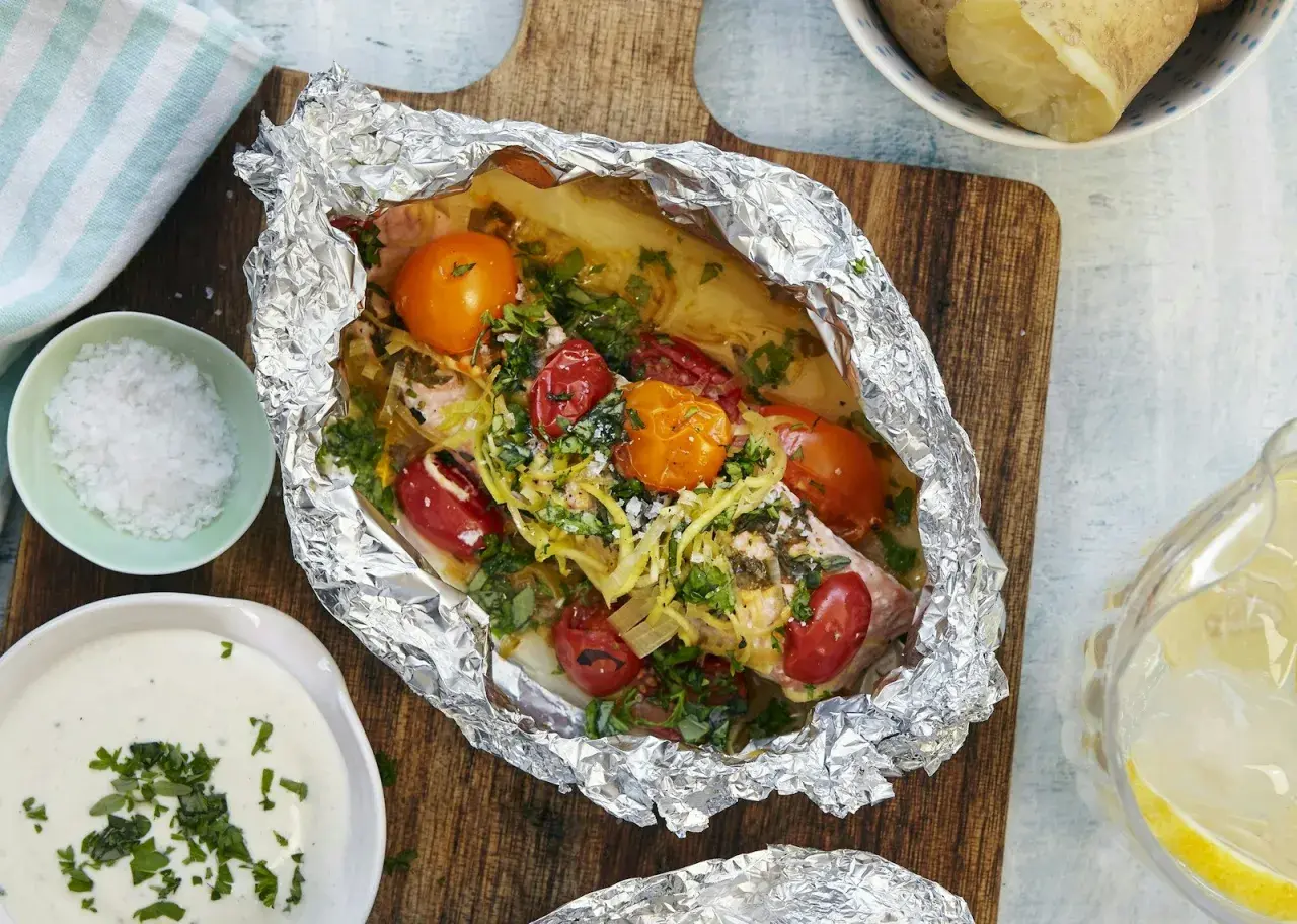 En saftig laxsida i foliepaket med tomater och örter, redo att grilla. Serveras med potatis och en krämig sås.
