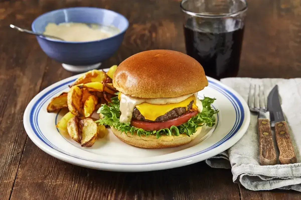 En saftig hamburgare med ost, sallad och tomat, serverad med klyftpotatis och en skål med krämig hamburgerdressing.